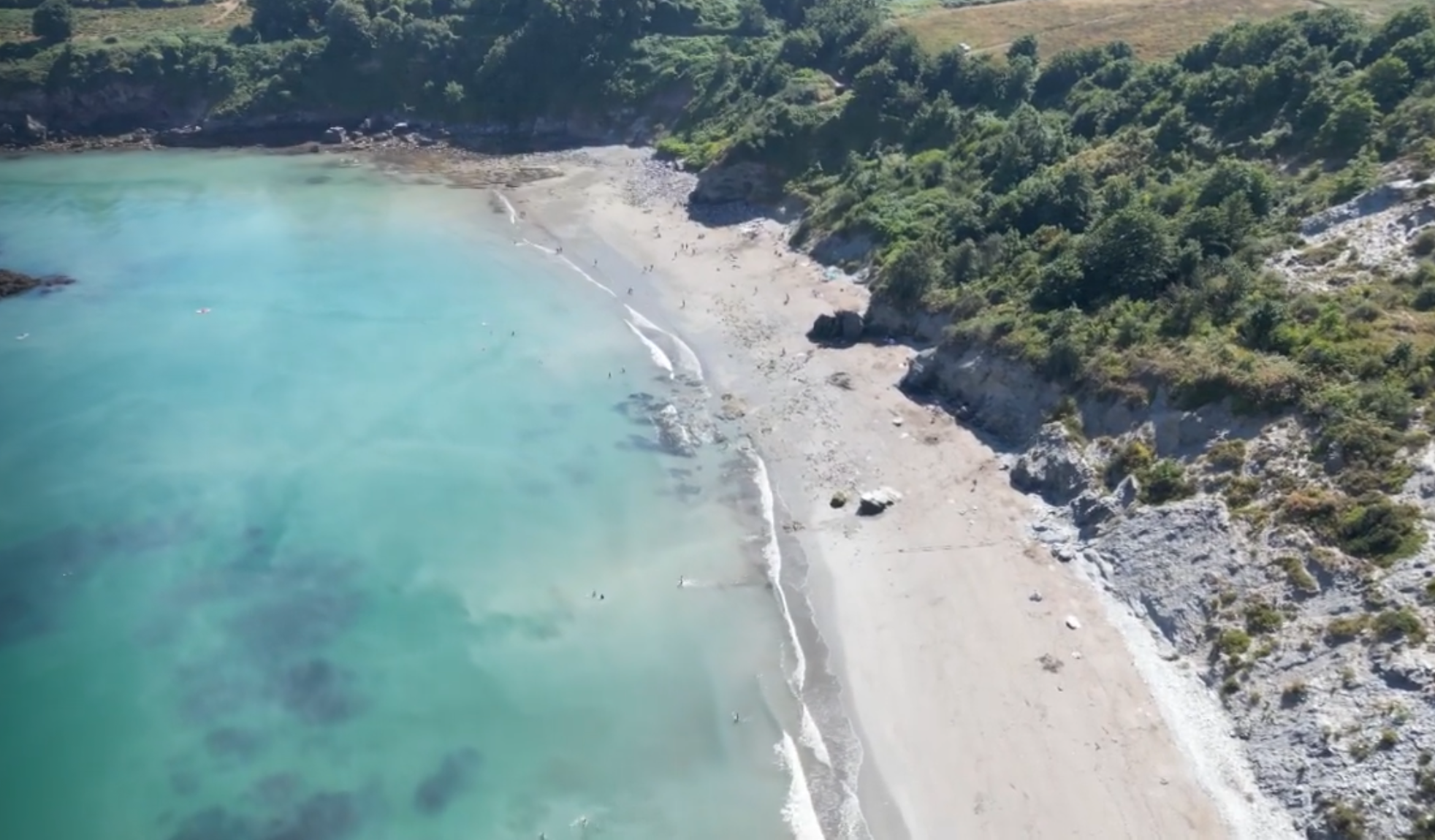 Aerial view of a scenic coastal cove with turquoise water and a sandy beach bordered by rocky cliffs and lush greenery. Small figures can be seen enjoying the beach and shallow waters, emphasizing the natural beauty and serenity of the location.